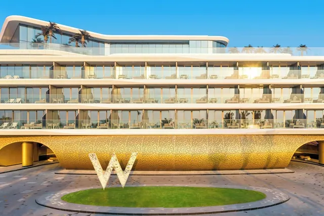 The exterior of a gold-clad hotel building with a large W logo sign and a green oval lawn on a paved forecourt, with loungers visible on the glazed balconies.