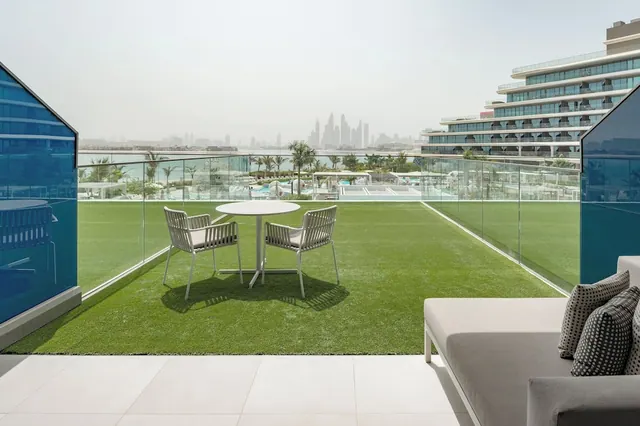 A furnished terrace with artificial grass, a round table with two chairs and a sofa, overlooking a pool, the sea and a distant city skyline.