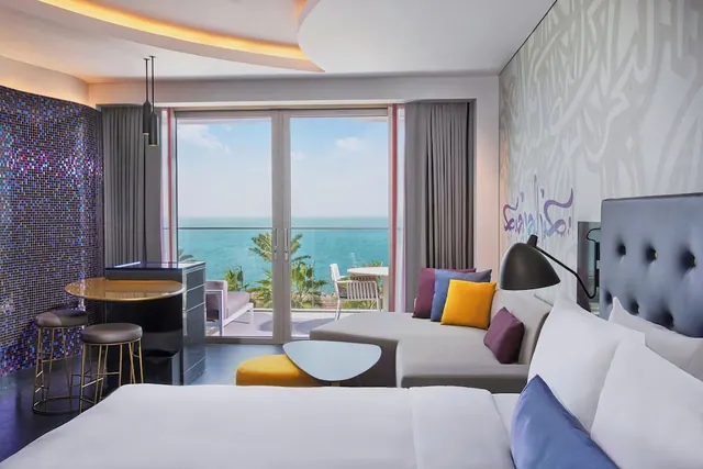 A hotel bedroom with a large bed, a grey sofa and a bar counter with stools, and glass doors opening onto a balcony with a view of the sea and palm trees.