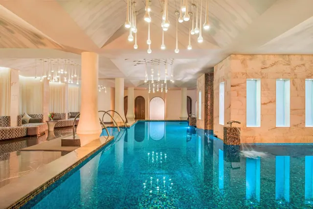 An indoor swimming pool with illuminated water, decorative pendant lights and lounge seating.