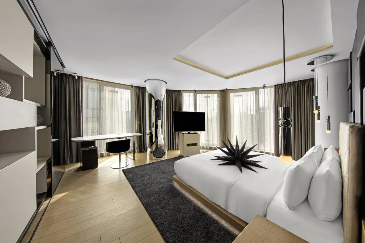 A bedroom with a large bed dressed in white linen, a round-edged desk and black chair, a television on a cabinet, and tall windows with sheer curtains.