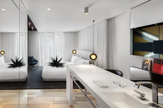 A hotel bedroom with two beds dressed in white linen and black star-shaped decorations, pendant lamps, a mirrored wall and a white counter with a sink and glasses.