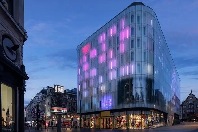 A curved glass-fronted building with pink and purple lighting stands at dusk beside older stone buildings and a street clock.