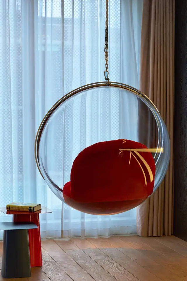 A transparent hanging bubble chair with a red cushion hangs in front of sheer white curtains, with a small red side table and a grey stool on the wooden floor beside it.