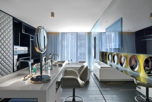 A modern bathroom with a long white vanity, a round illuminated mirror, a white bathtub by a sheer-curtained window, and grey swivel stools.