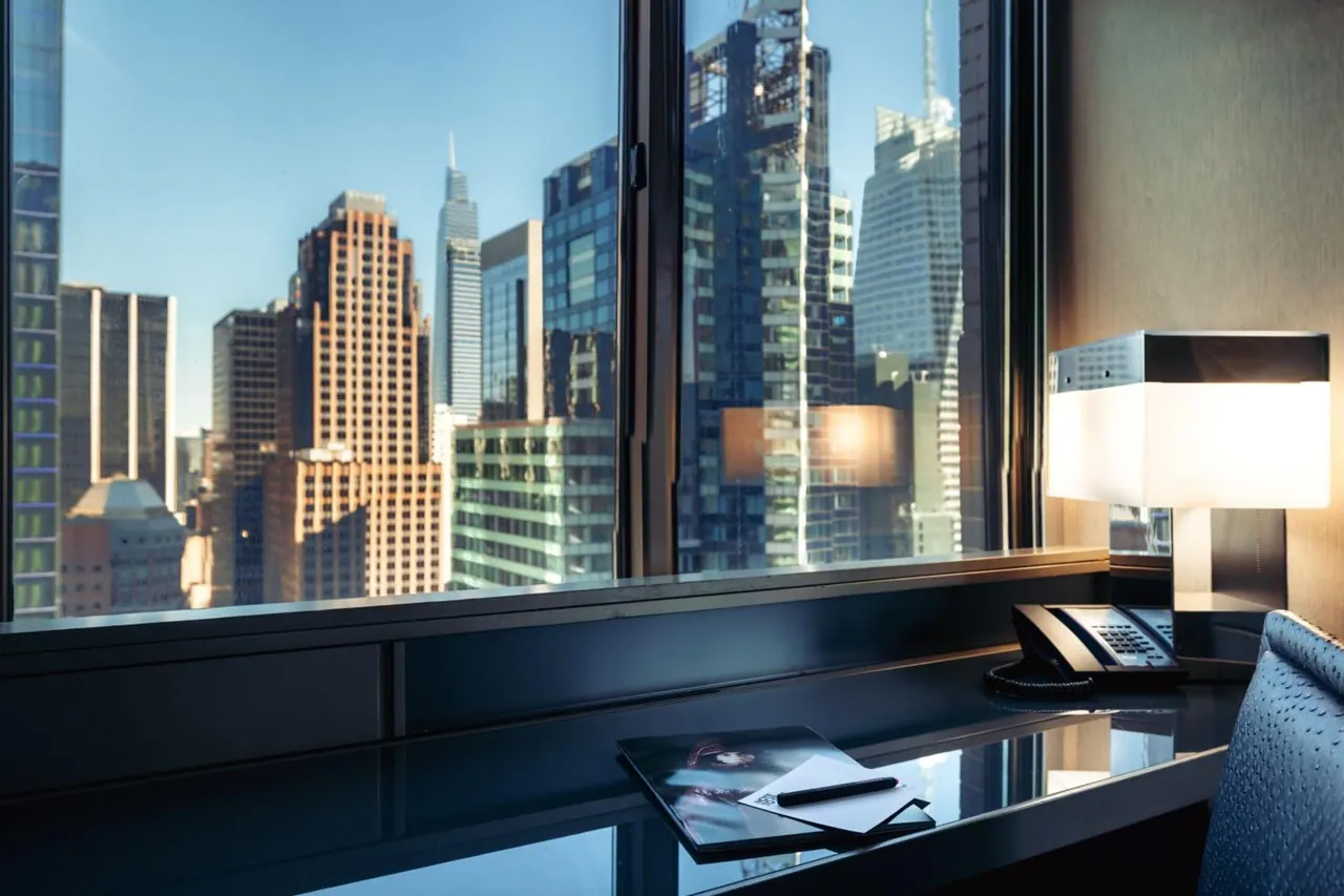 A desk by a window looks out on a city skyline of tall buildings, with a telephone, a lamp and a folder with a pen on the desk.