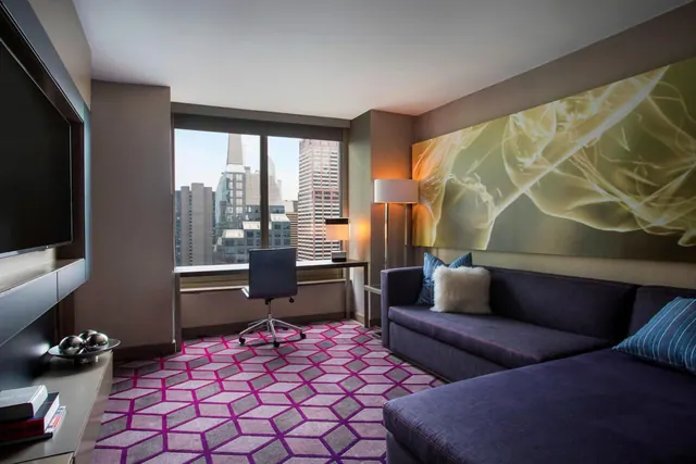 A hotel room with a purple sectional sofa, a desk and chair by a window looking out over city buildings, and a wall-mounted television on the left.