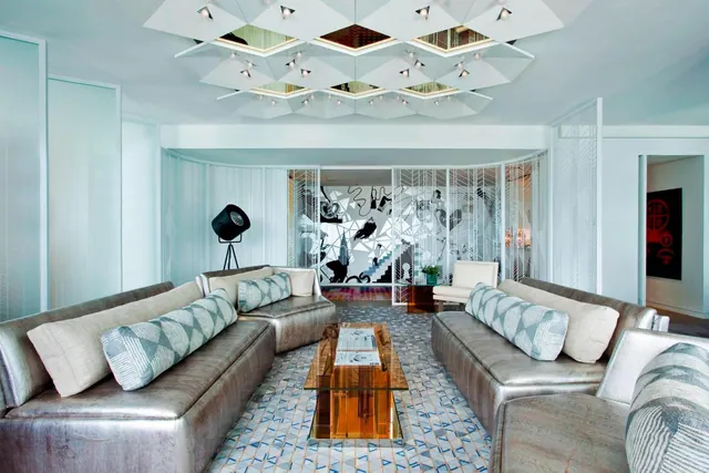 A living area with two metallic grey sofas facing a glass coffee table, under a geometric white ceiling with spotlights and a black-and-white mural on the back wall.