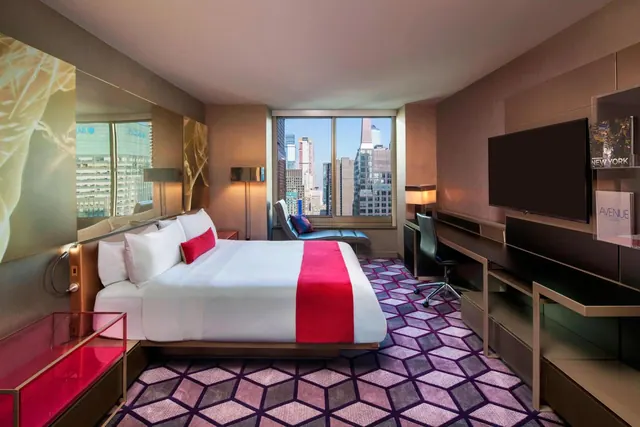 A king bed with a red runner and white pillows sits in a hotel room beside a desk, a wall-mounted television and a window looking out over city skyscrapers.