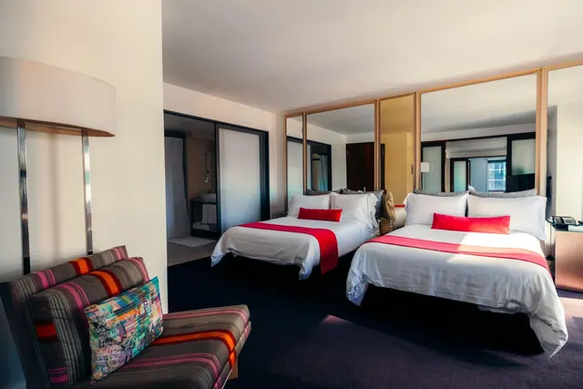 A hotel bedroom with two double beds dressed in white linen and red runners, a striped armchair in the foreground, mirrored wardrobe doors, and an open doorway to the bathroom.
