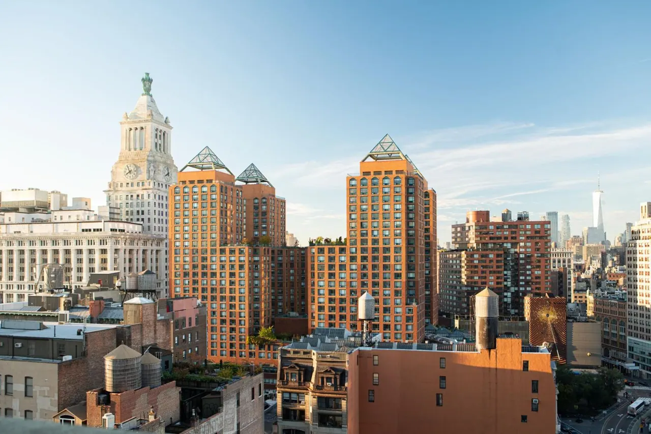 A view over brick towers, a white clock tower and the distant One World Trade Center under a clear sky.