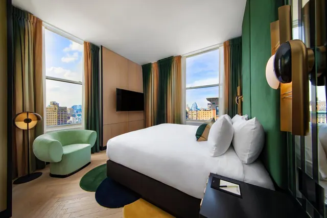 A hotel bedroom with a king bed, a mint green armchair, a wall-mounted television and two windows looking out over city buildings.