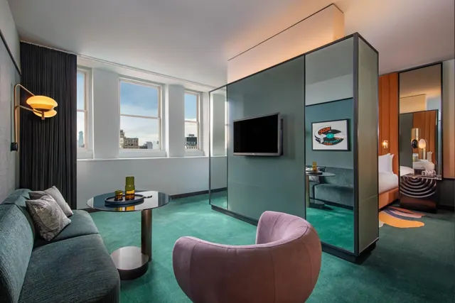 A living area with a teal sofa, purple armchair, round side table and wall-mounted television, with city buildings visible through the windows and a bed seen beyond a mirrored partition.