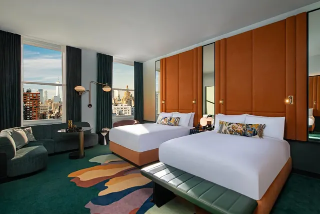 A hotel bedroom with two beds that have orange upholstered headboards, a grey sofa, and tall windows overlooking a city skyline that includes One World Trade Center.
