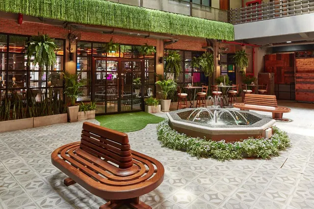 An indoor atrium with wooden benches, a patterned tiled floor, a fountain ringed by white flowering plants, and a restaurant entrance with glass doors behind.