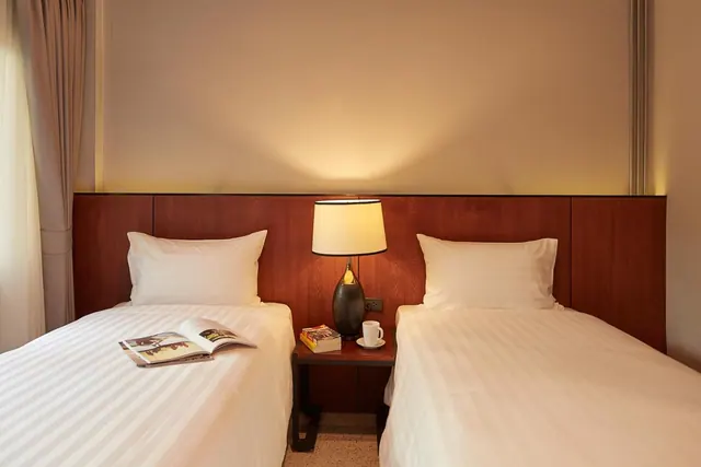 Two single beds with white striped linen and a wooden headboard, with a bedside lamp, a cup and books on the nightstand, and an open magazine on the left bed.