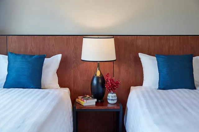 Two twin beds with blue cushions and white striped linen sit either side of a wood-panelled headboard and a bedside table holding a lamp, books and a vase of red flowers.