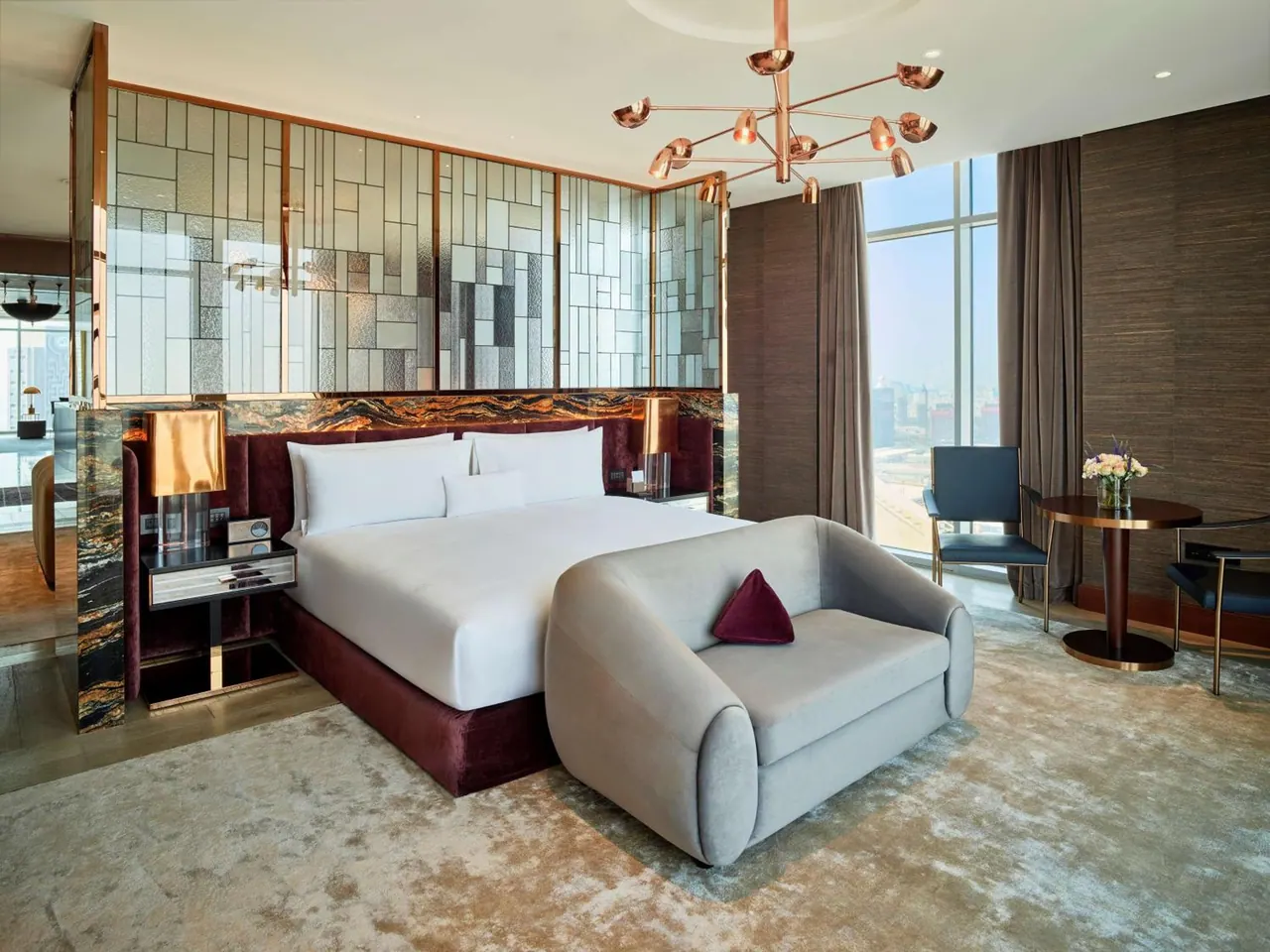 A bedroom with a made king bed, a grey armchair and a round side table, with floor-to-ceiling windows showing a hazy city skyline.