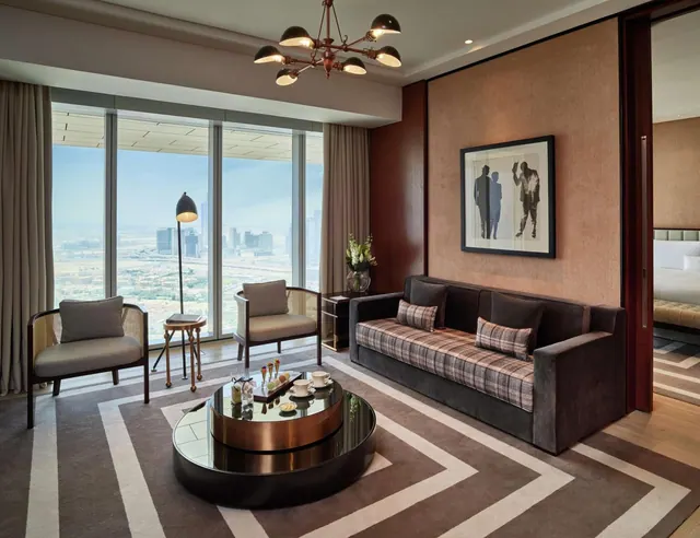A living area with a sofa, two armchairs, a round coffee table and floor-to-ceiling windows overlooking a city skyline, with a bed visible through an open doorway.