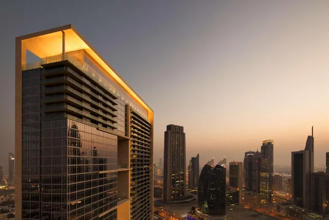 A dusk exterior view of a tall glass tower with an illuminated overhanging top, with other city towers and lights visible behind it.