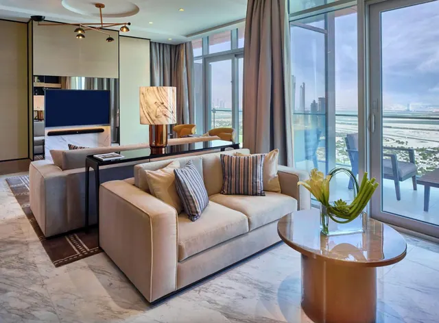 A living area with two beige sofas, a round wooden coffee table holding a vase of flowers, a wall-mounted television, and sliding glass doors opening onto a balcony with a view of distant towers and open land.