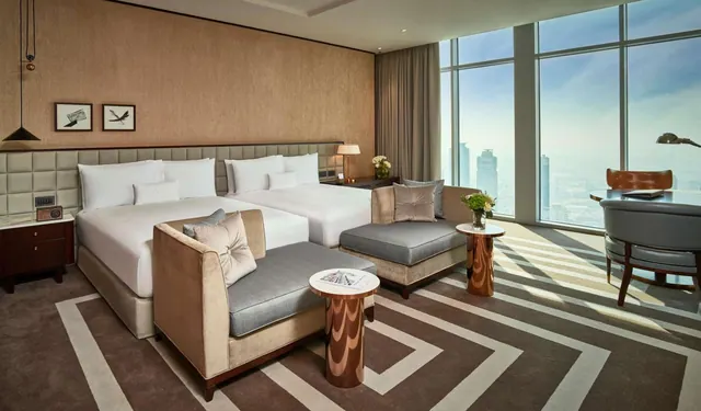 A bedroom with two made beds, two armchairs with side tables, a desk and chair, and floor-to-ceiling windows looking over a hazy city with tall towers.