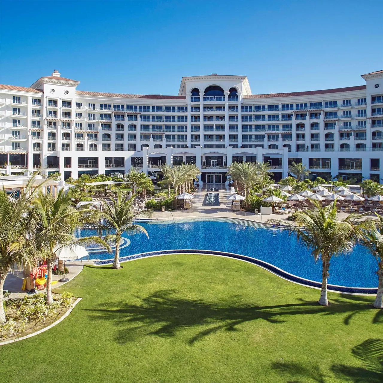 A large curved swimming pool with lounge chairs and white umbrellas sits in front of a white multi-storey hotel building with palm trees and a green lawn.