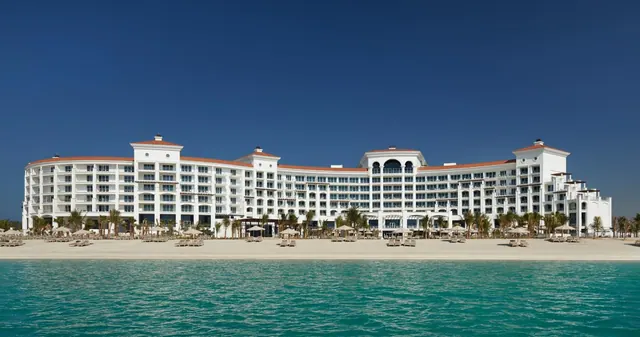 A large white multi-storey hotel building stands behind a sandy beach with sun loungers and umbrellas, facing turquoise water under a clear blue sky.