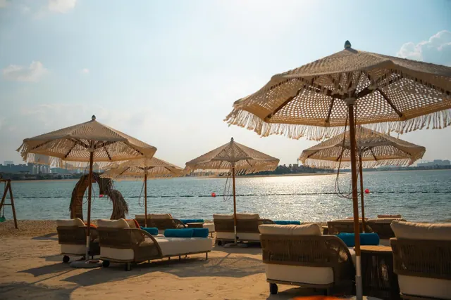 Woven fringed parasols and cushioned sun loungers with blue bolsters stand on sand facing a calm sea.