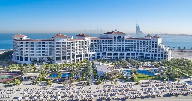 Aerial view of a large white beachfront hotel with pools, palm trees and rows of loungers on the sand, with the Burj Al Arab visible in the distance.