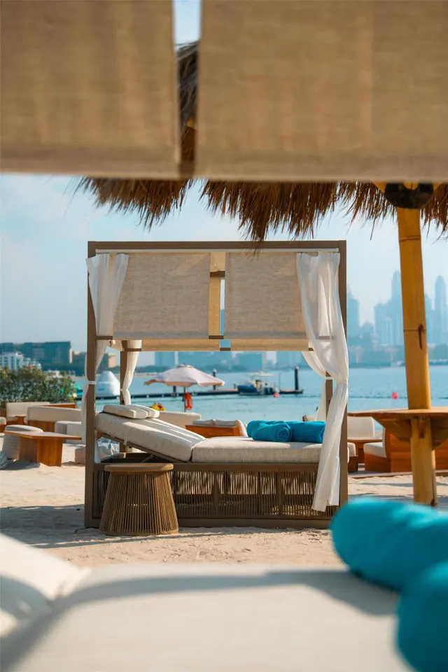 A canopied daybed with teal cushions stands on sand under a thatched shade structure, with the sea and a city skyline of towers beyond.