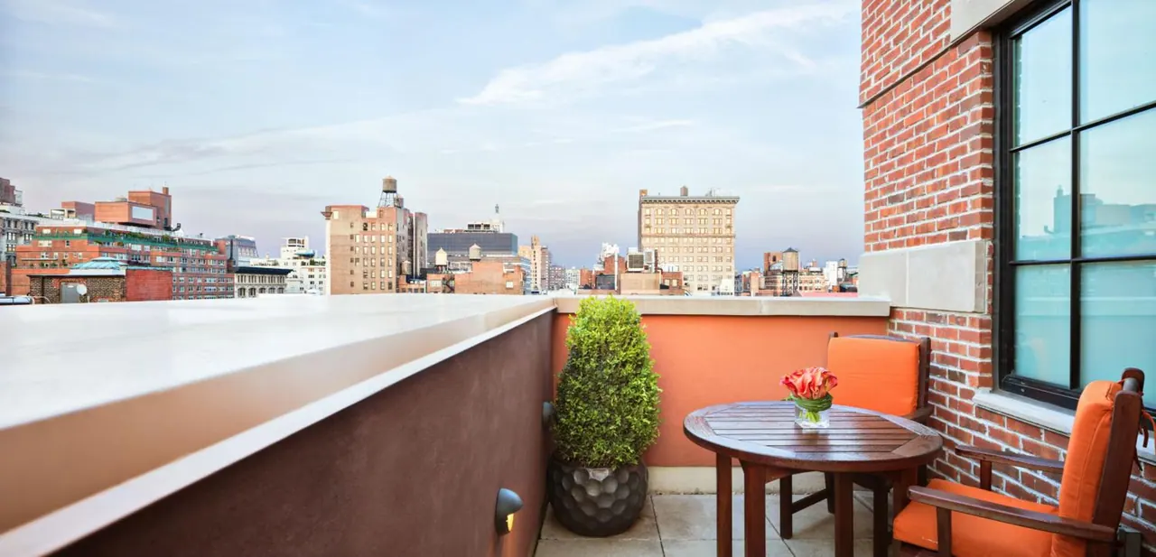 A rooftop terrace with orange cushioned chairs, a round wooden table with tulips, a potted topiary and a view over city buildings.