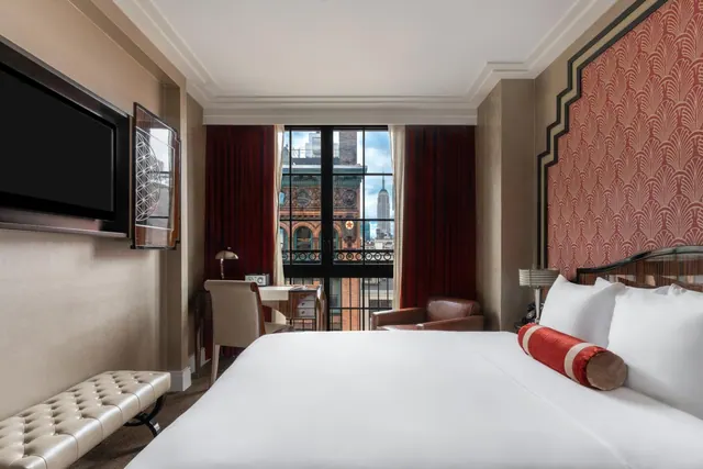 A made king bed with a red bolster sits in a room with a television, a desk, a leather armchair and a window showing a brick building and the Empire State Building.
