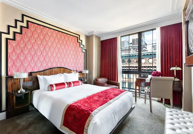 A king bed with a red and white throw sits against a patterned art deco headboard wall, with a desk, armchair and a window looking onto a building facade.