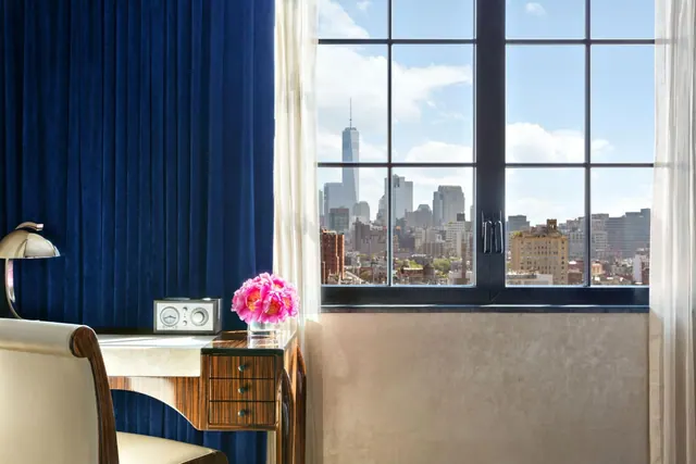 A wooden desk with a vintage radio, a lamp and a vase of pink peonies sits beside a window overlooking a city skyline that includes One World Trade Center.