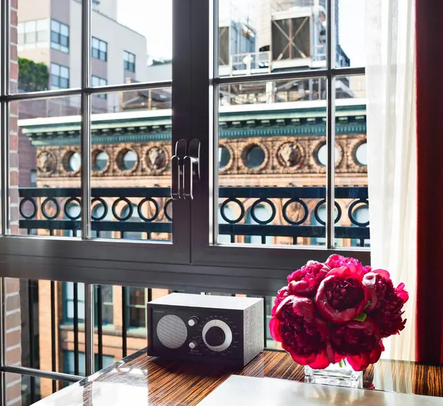 A window looks out onto neighbouring city buildings and an ornate cast-iron balcony, with a radio and a vase of red peonies on a wooden table in front.