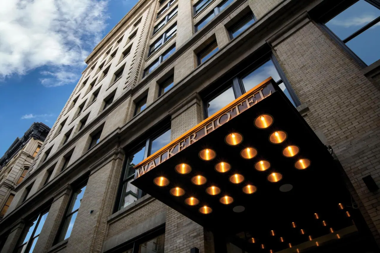 Looking up at the brick facade of a hotel building, with a lit canopy sign reading WALKER HOTEL under a partly cloudy blue sky.