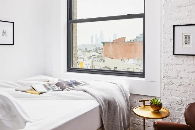 A made bed with white linens, an open magazine and a grey throw sits beside a window looking over rooftops and distant towers.