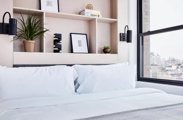 A made bed with white pillows sits beside a built-in shelving unit and a window looking out over city buildings.