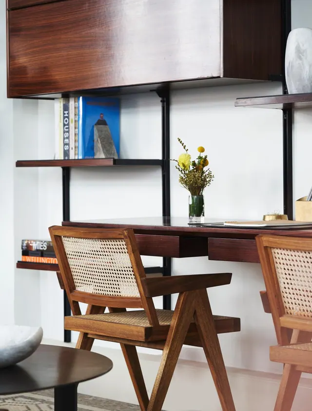A wooden desk with a vase of flowers sits beneath a wall-mounted cabinet and shelves of books, with cane-backed chairs in front.