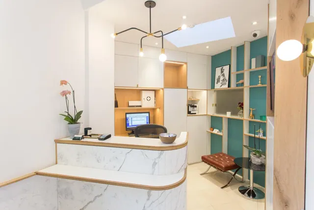 A reception counter with a white marble front stands beside a computer monitor, a leather stool, a potted orchid and built-in shelving against a teal wall, under a skylight.