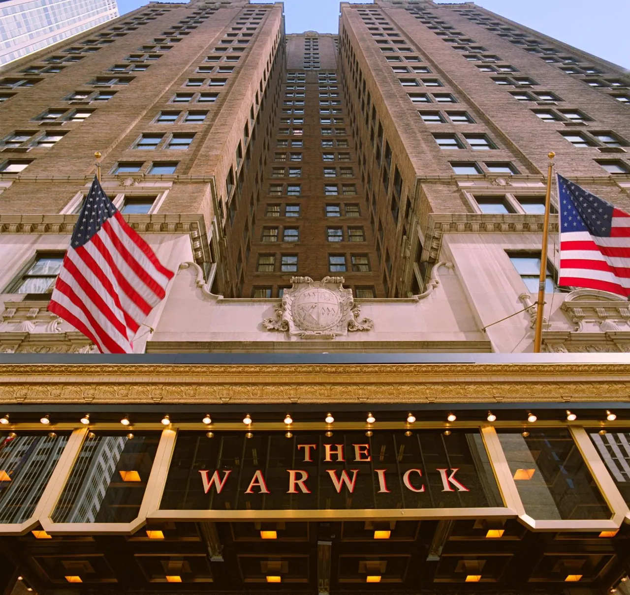 The exterior of The Warwick hotel tower is shown from street level, with American flags on poles and the name sign on the entrance canopy.