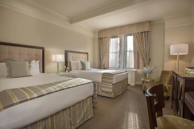 A bedroom with two made beds with tufted headboards, a window with draped curtains, an armchair and a wooden desk with a chair.