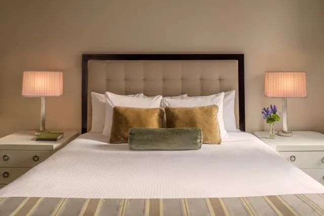 A made king bed with a tufted headboard and decorative pillows, flanked by two lit table lamps on white nightstands, with a small flower vase on the right nightstand.