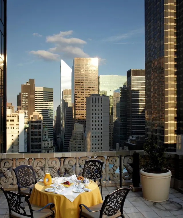 A set breakfast table with a yellow tablecloth and metal chairs sits on a stone terrace overlooking a city skyline of tall office towers under a blue sky.