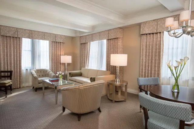 A hotel suite living area with a cream sofa, armchairs, a glass coffee table, two table lamps and sheer-curtained windows.
