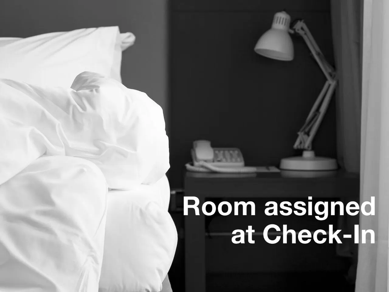 A made bed with white linens sits beside a dark nightstand holding a telephone and an angled desk lamp, in black and white, with a text overlay reading Room assigned at Check-In.