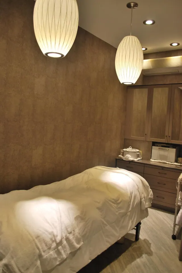 A treatment room with a massage table covered in white linens, two pendant lamps, wood-fronted cabinets and a dresser holding a towel warmer.