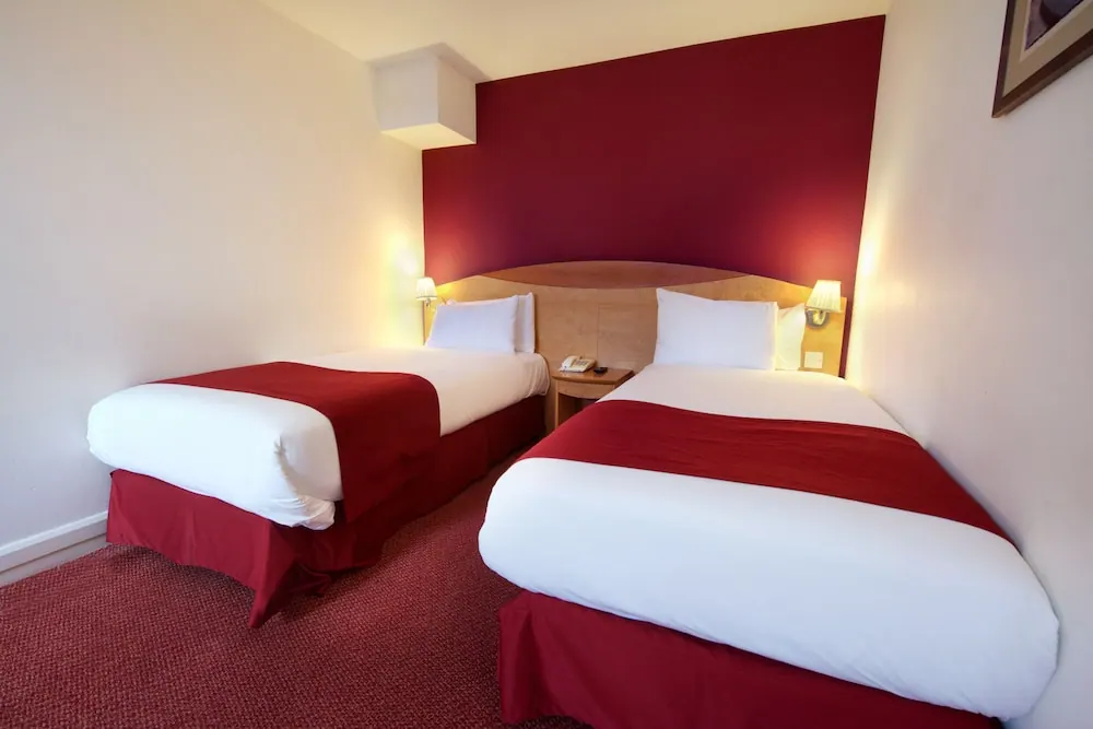 A hotel bedroom with two single beds with white bedding and red runners, a red accent wall and red carpet.
