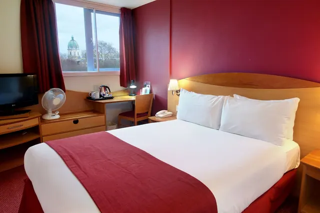 A double bed with a red headboard wall sits beside a desk with a kettle tray, a television on a cabinet, and a window showing a green-domed building and trees outside.